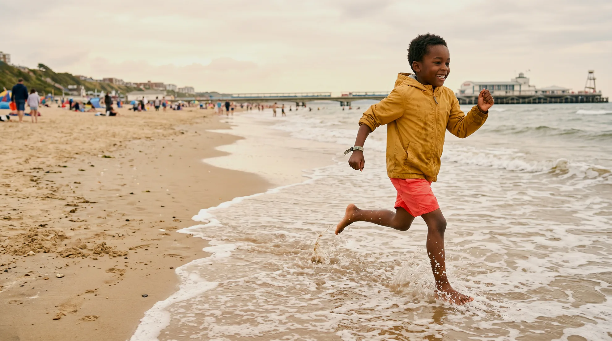 A young boy in a mustard jacket and coral shorts laughing as he runs through the waves on Bournemouth beach, with the pier in the background