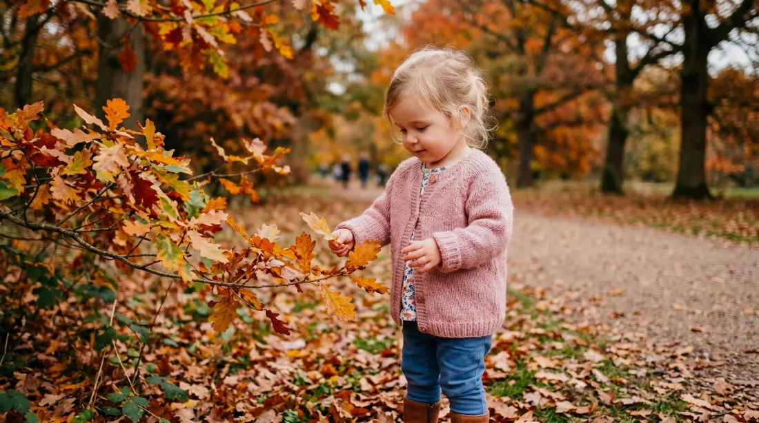 A toddler in a pink knitted cardigan picks up an autumn leaf on a leaf-covered woodland path